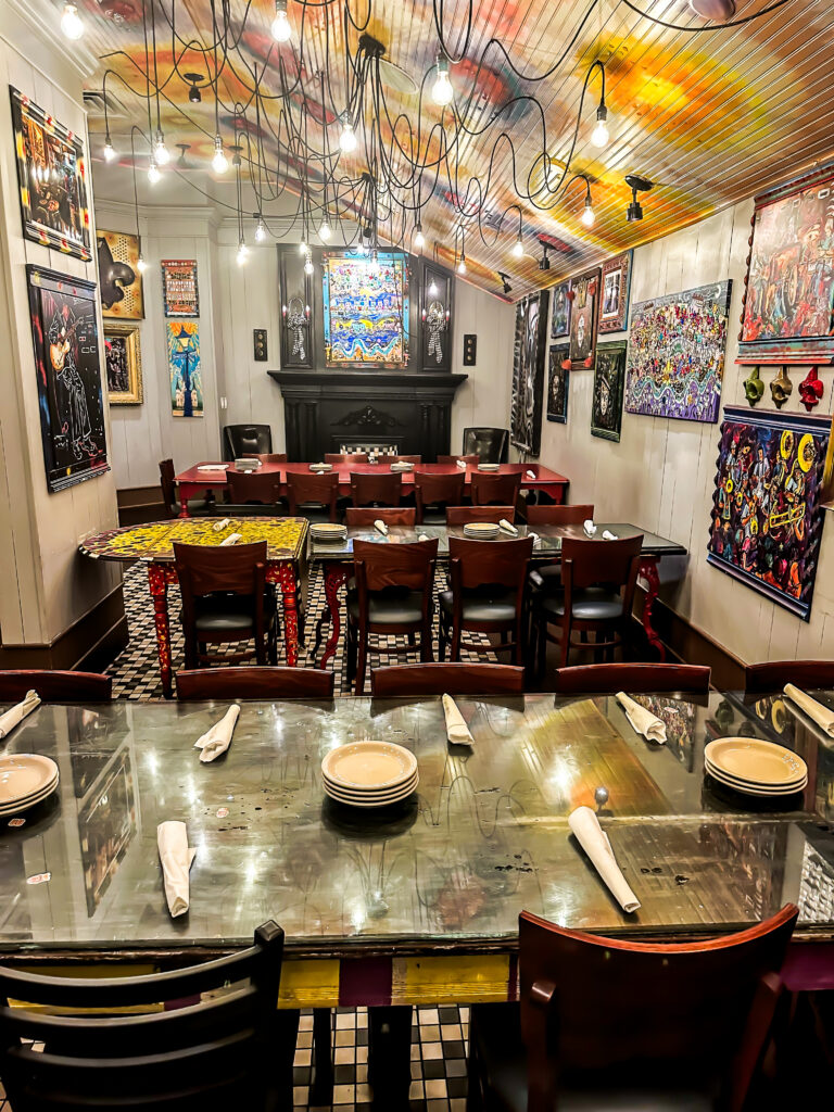 A wide view of the Napoleon Room at Tibby’s Brandon, tables set beneath a painted ceiling hung with bulbs, walls covered in framed New Orleans folk art