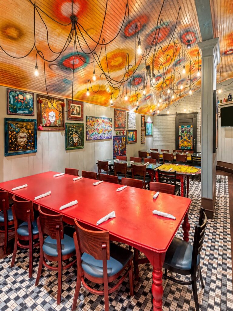 The Napoleon Room at Tibby’s Brandon, its long red table set beneath a hand-painted ceiling strung with bulbs, walls covered in New Orleans folk art